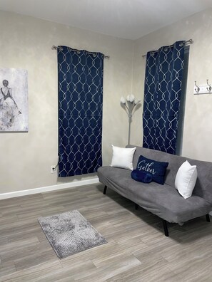 Een woonkamer met een grijze bank, een blauw gordijn met patroon, een grijze vloerkleed en een wandlamp.