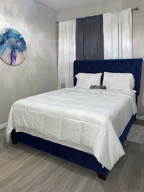 Een bed met een witte quilt, een blauw hoofdbord en een grijze kussen.