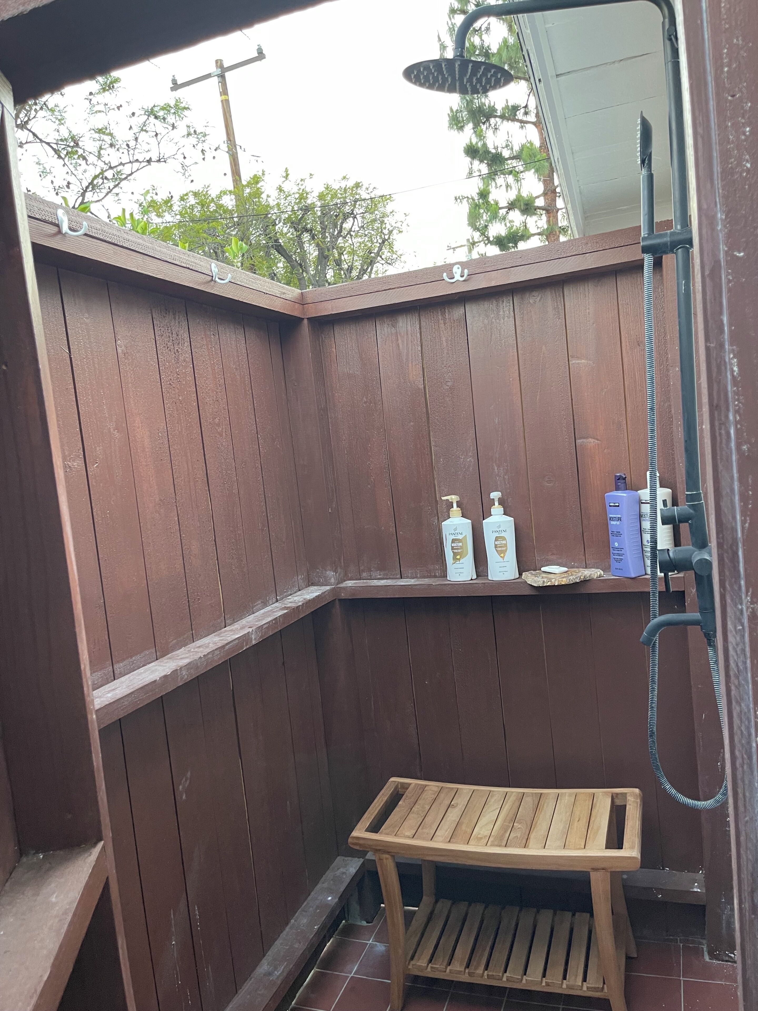 Un área de ducha exterior de madera con un banco, una manguera y dos botellas de champú Pantene.