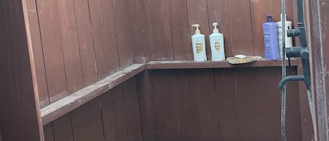 Un espace de douche extérieur en bois, avec un banc, un tuyau d’arrosage et deux bouteilles de shampoing Pantene.