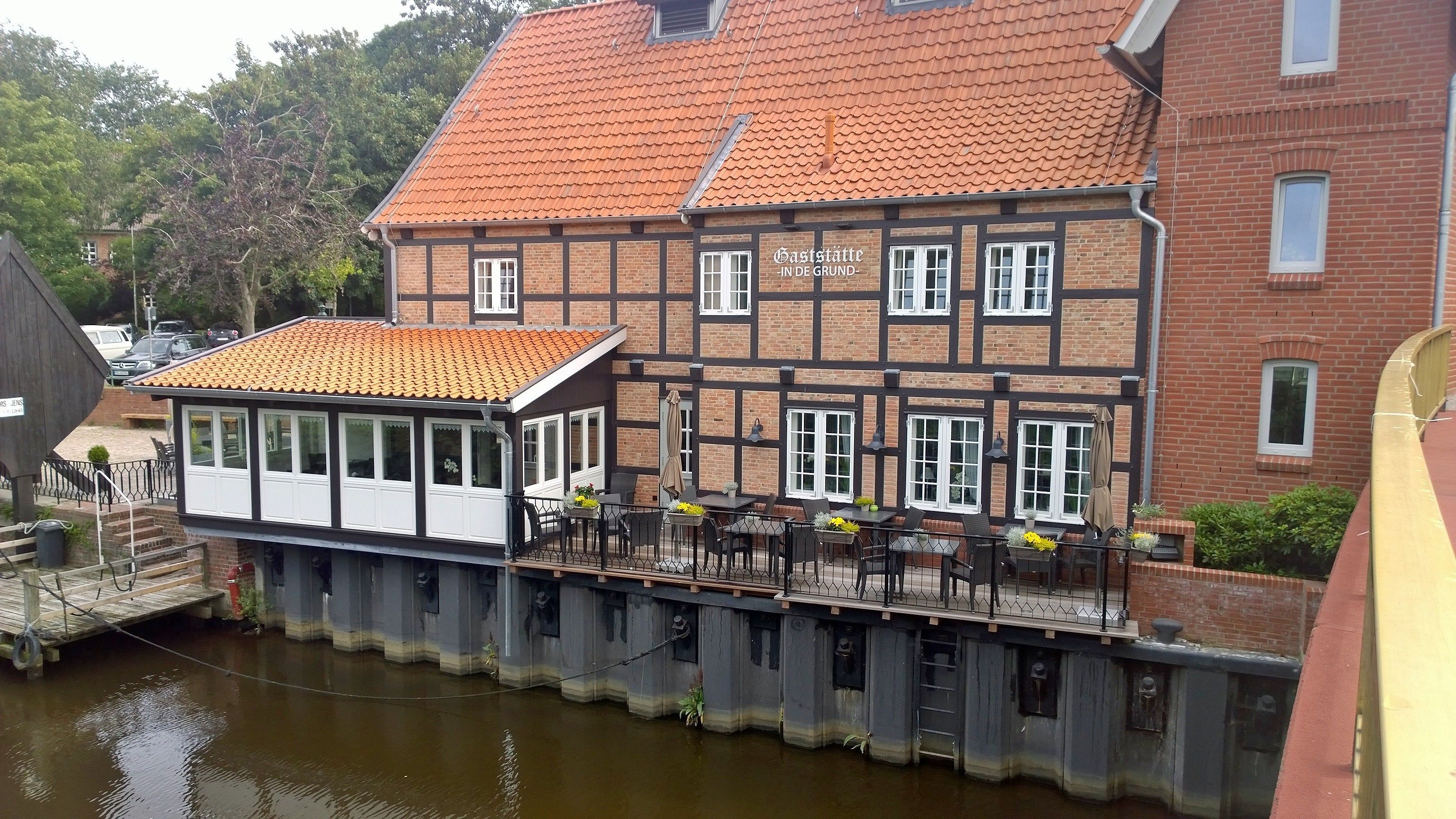 A traditional half-timbered building with a red-tiled roof, a covered outdoor seating area with tables and chairs, and a wooden dock extending over a body of water.