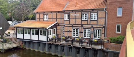 En traditionel bindingsværksbygning med rødt tegltag, et overdækket udendørs siddeområde med borde og stole, og en træbro, der strækker sig ud over vandet.