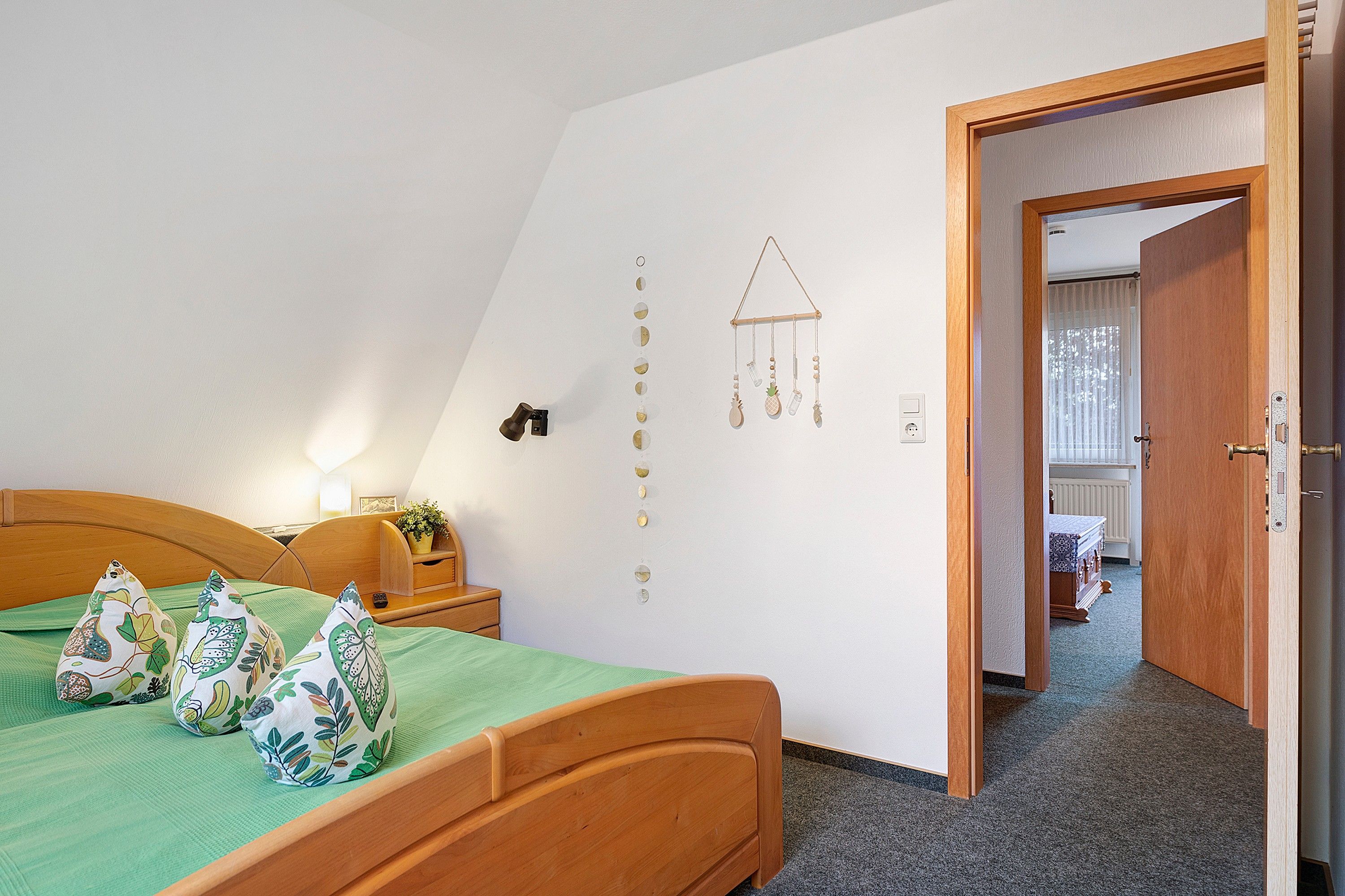 A bedroom with a wooden bed, a bedside table with a lamp, a wall decoration, and a door leading to another room.