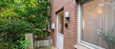 A brick building with a large window, a door, and a small outdoor area with a wooden fence and greenery.