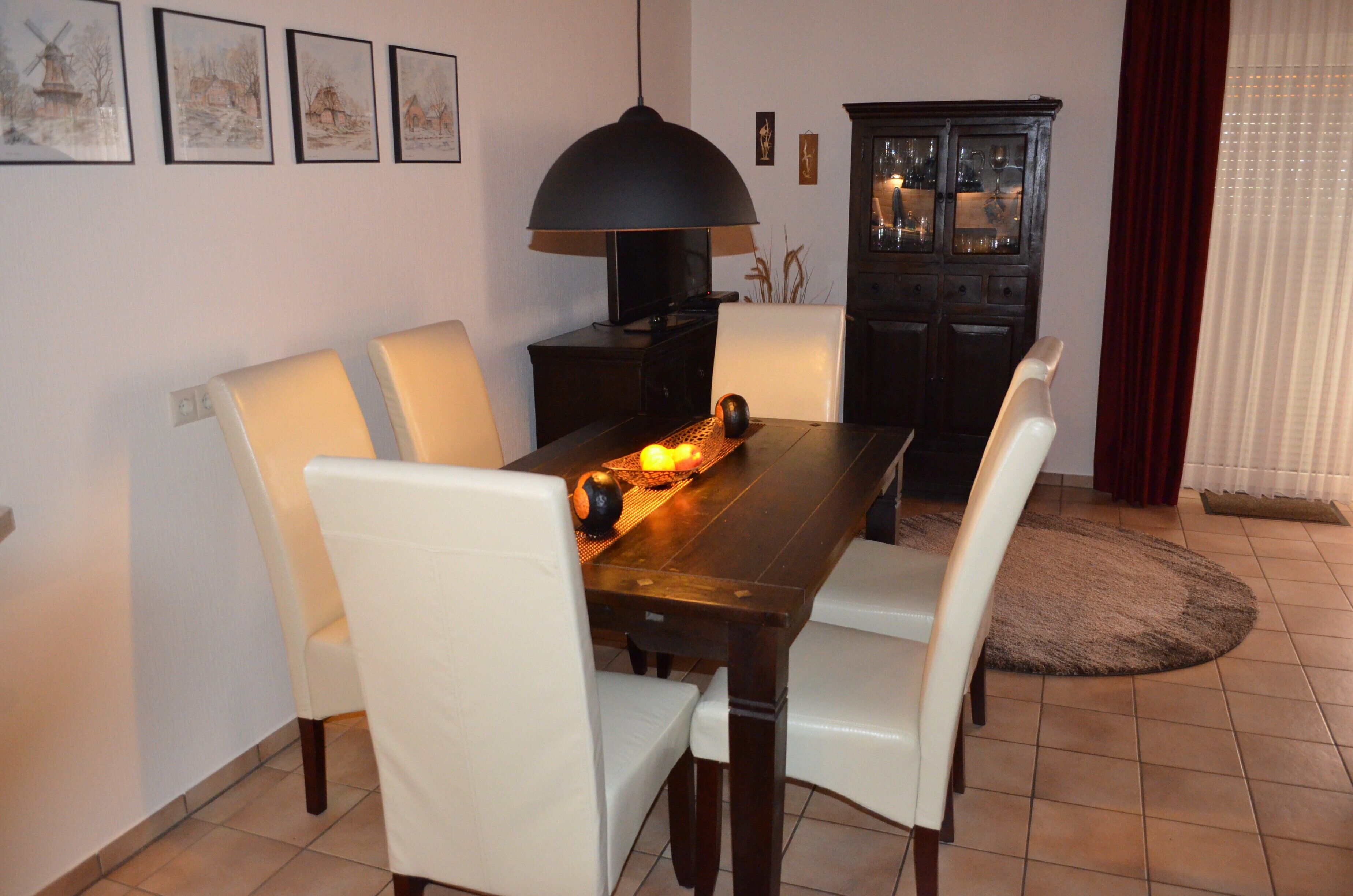 Zona pranzo con tavolo in legno, sedie bianche, lampadario nero e un armadietto con ante in vetro.