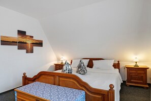 A bedroom with a wooden bed, two bedside tables with lamps, a nightstand, a patterned bench, and a wall with a sunset painting. - Holiday Home Godewind with Terrace, Garden, Wi-Fi & Balcony Near Beach in Otterndorf (Otterndorf)