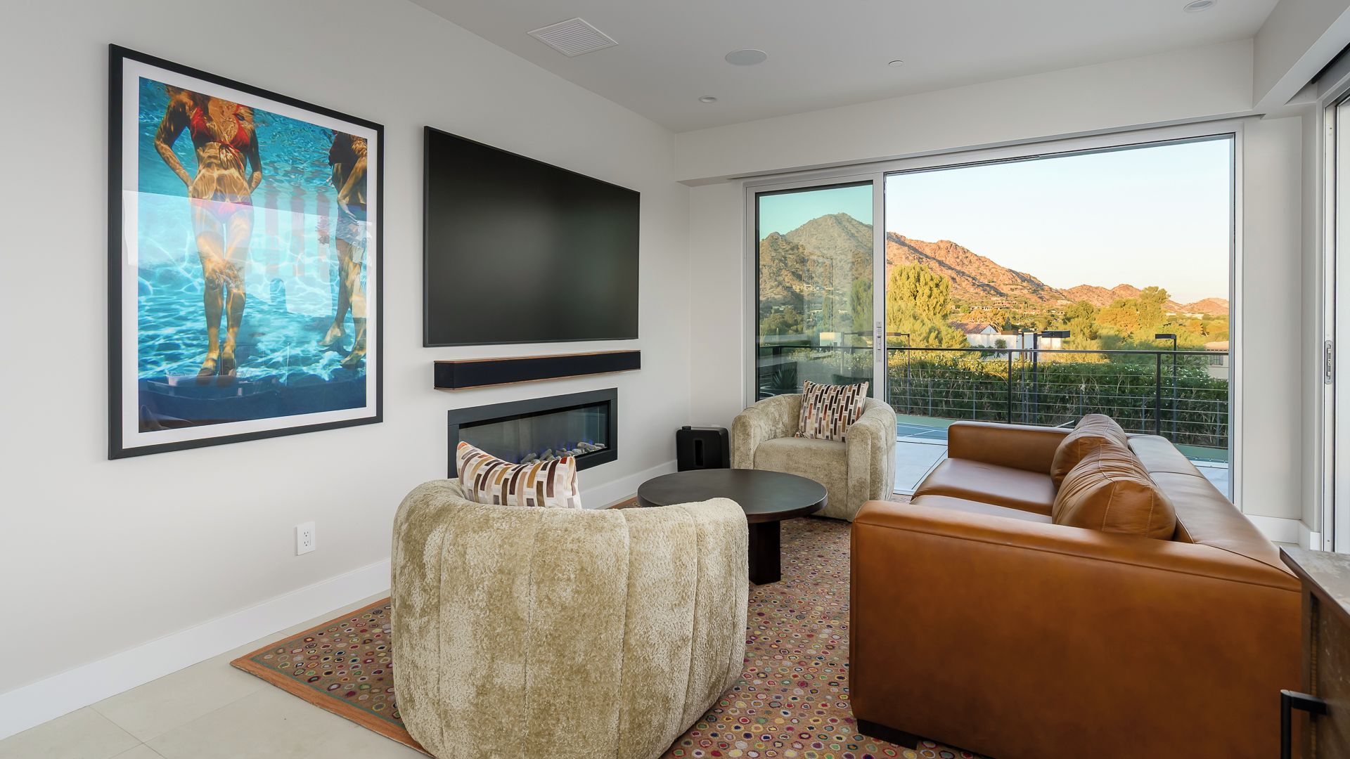 A modern living room with a large flat-screen TV, comfortable seating, a fireplace, and a balcony with a mountain view.