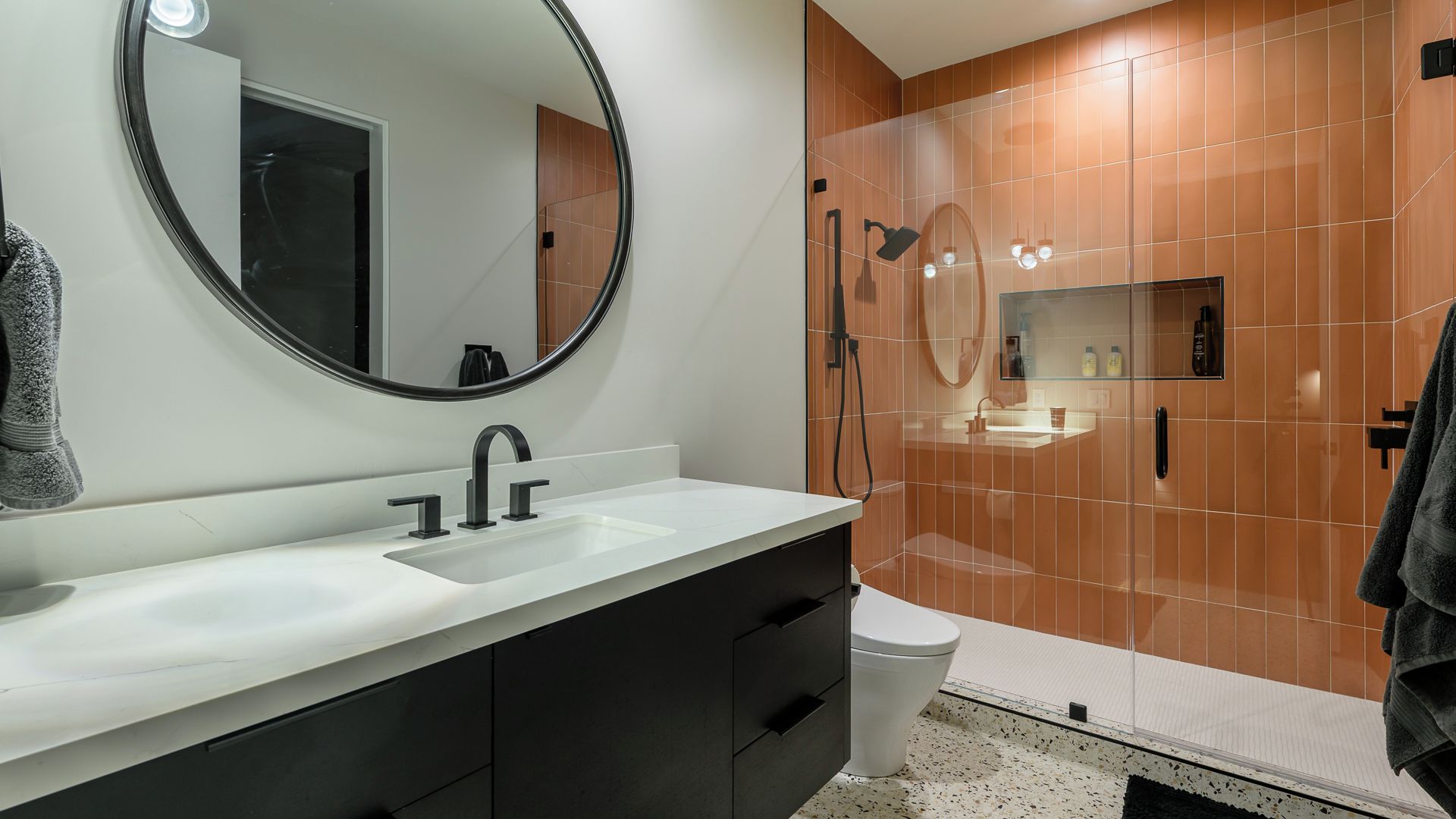 Un bagno moderno con un lavandino bianco, mobili neri, uno specchio rotondo e un box doccia in vetro.