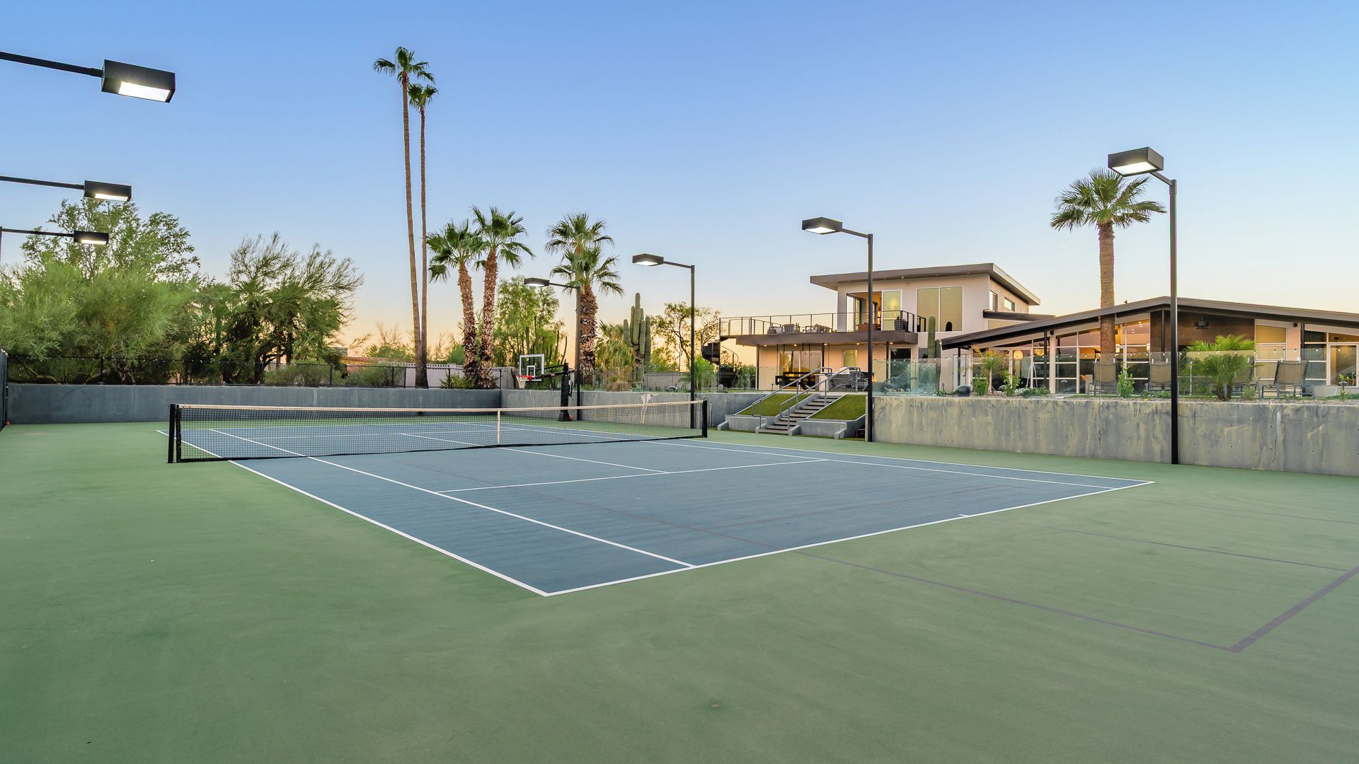 Un terrain de tennis avec un filet, entouré de palmiers et éclairé par des lampadaires, avec un bâtiment en arrière-plan.