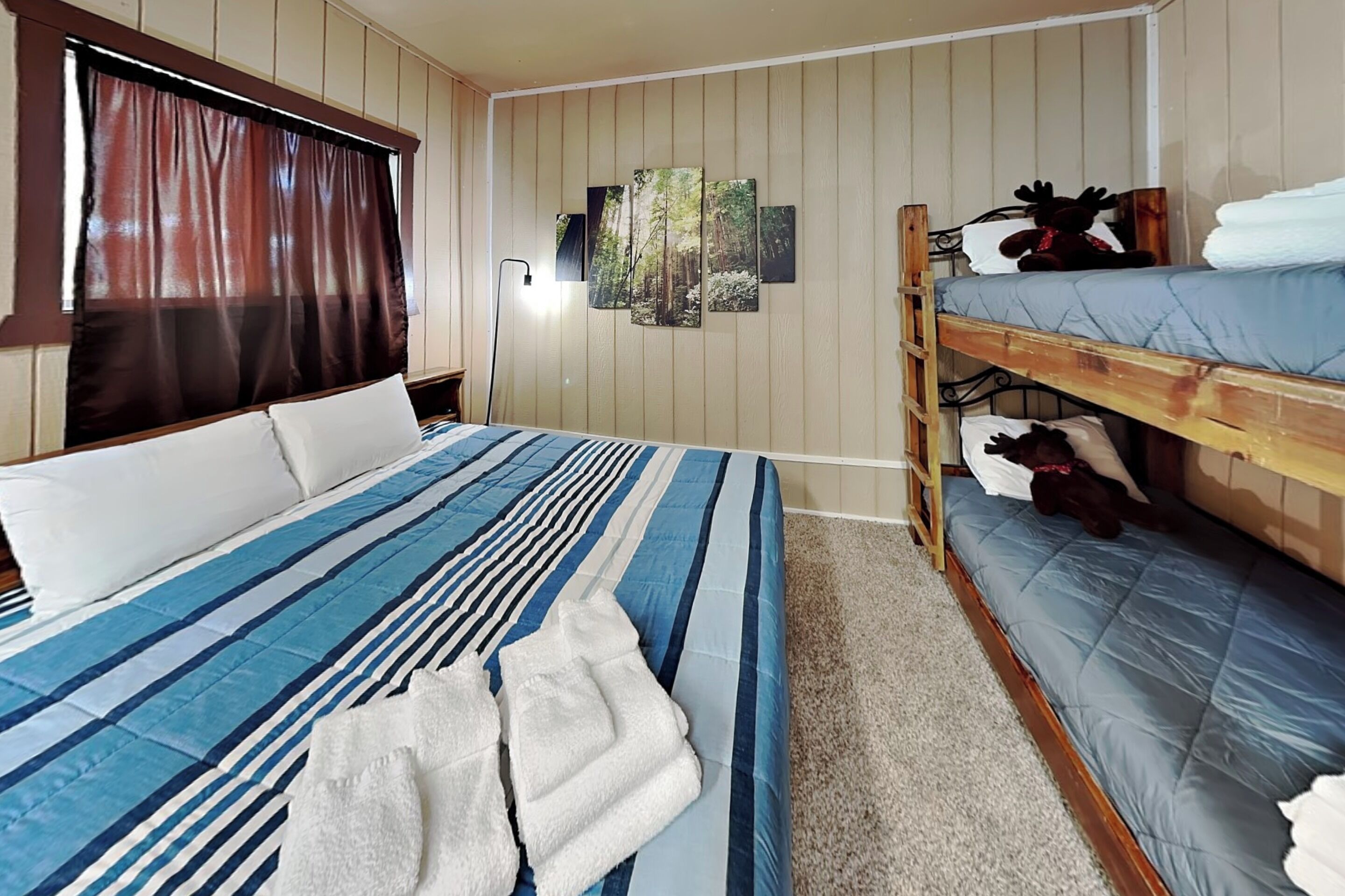 A room with a bunk bed, a large bed with striped bedding, a window with curtains, and a wall with framed pictures.
