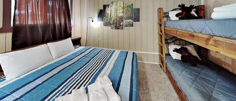 A room with a bunk bed, a large bed with striped bedding, a window with curtains, and a wall with framed pictures.