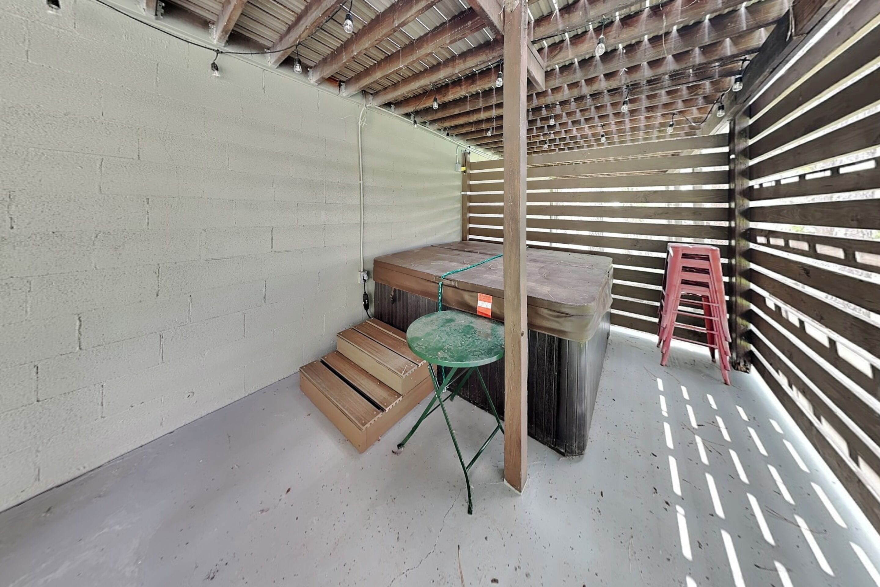 A covered outdoor area with a wooden bench, a small table, and a red stool.