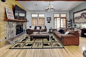 A cozy living room with a stone fireplace, a chandelier, and a large window with curtains. - Cozy Cove Lodge (Ruidoso)