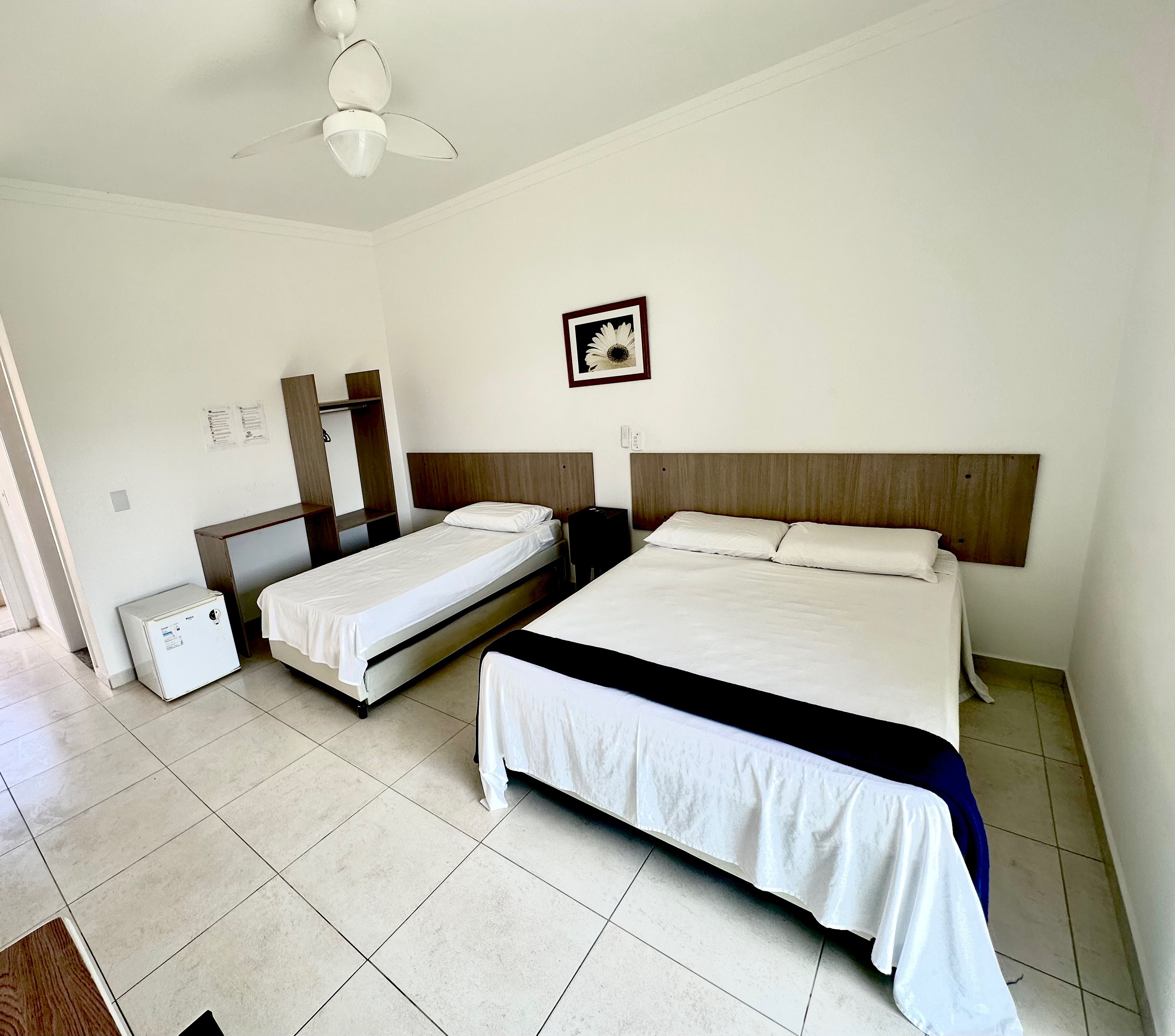 A hotel room with two single beds, a large bed, a ceiling fan, a small refrigerator, and a framed picture on the wall.