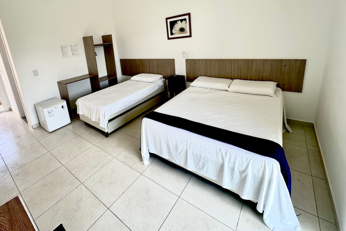 A hotel room with two single beds, a large bed, a ceiling fan, a small refrigerator, and a framed picture on the wall.