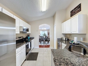 A kitchen with white cabinets, a stainless steel refrigerator, a granite countertop, and a view into the dining area. - Emerald Island Four Bed Pool Home close to Disney (Kissimmee)