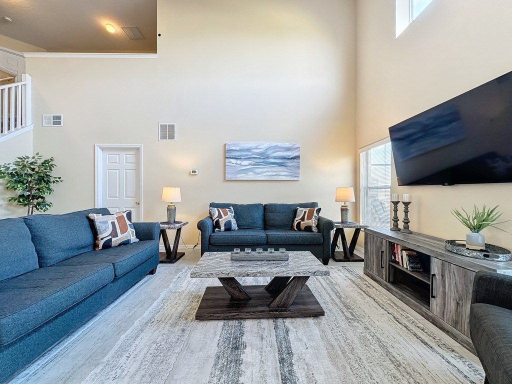 A spacious living room with a high ceiling, a large flat-screen TV mounted on the wall, a blue sofa set, a coffee table, and a wooden cabinet with books.