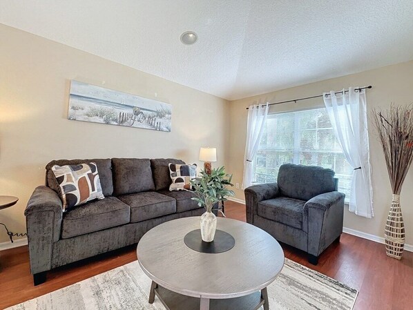 A living room with two grey sofas, a round coffee table, a painting on the wall, a window with curtains, and a vase with branches.