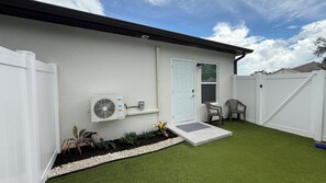 A residential property with a white exterior, a white door, a small patio area with artificial grass, a white fence, and an air conditioning unit mounted on the wall.