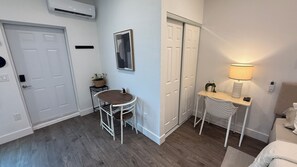 A room with a wooden floor, a round table with chairs, a small side table with a lamp, a potted plant, and a door.