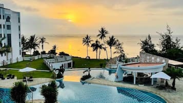 Zona de piscina en un resort con vista al océano al atardecer, palmeras y un edificio blanco.