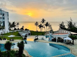 Zona de piscina en un resort con vista al océano al atardecer, palmeras y un edificio blanco.