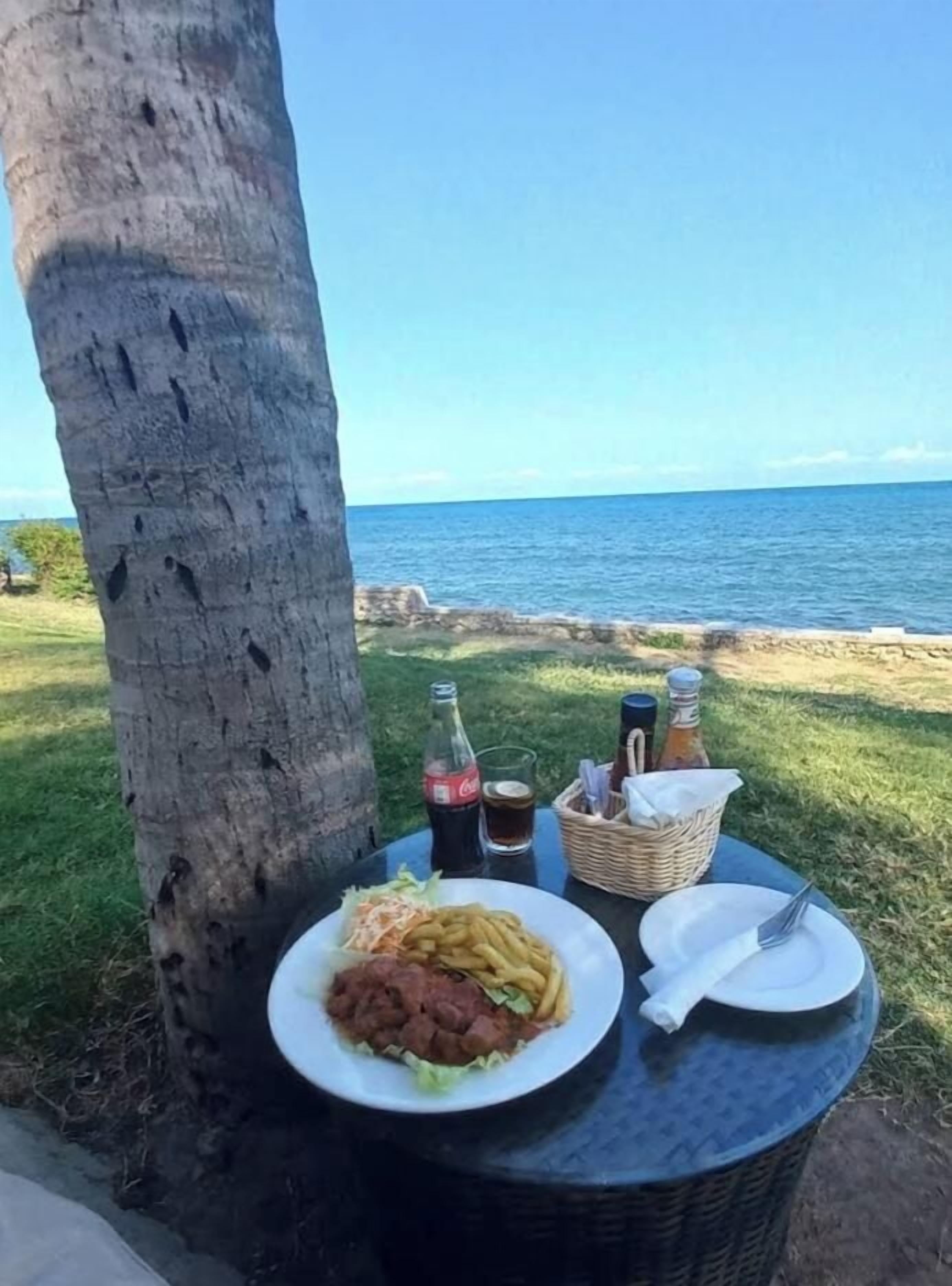 Una mesa con comida y bebidas, una botella de Coca-Cola y una cesta, ubicada al aire libre con vista al mar.