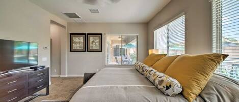 A bedroom with a bed, a TV, a ceiling fan, and a view through a window.