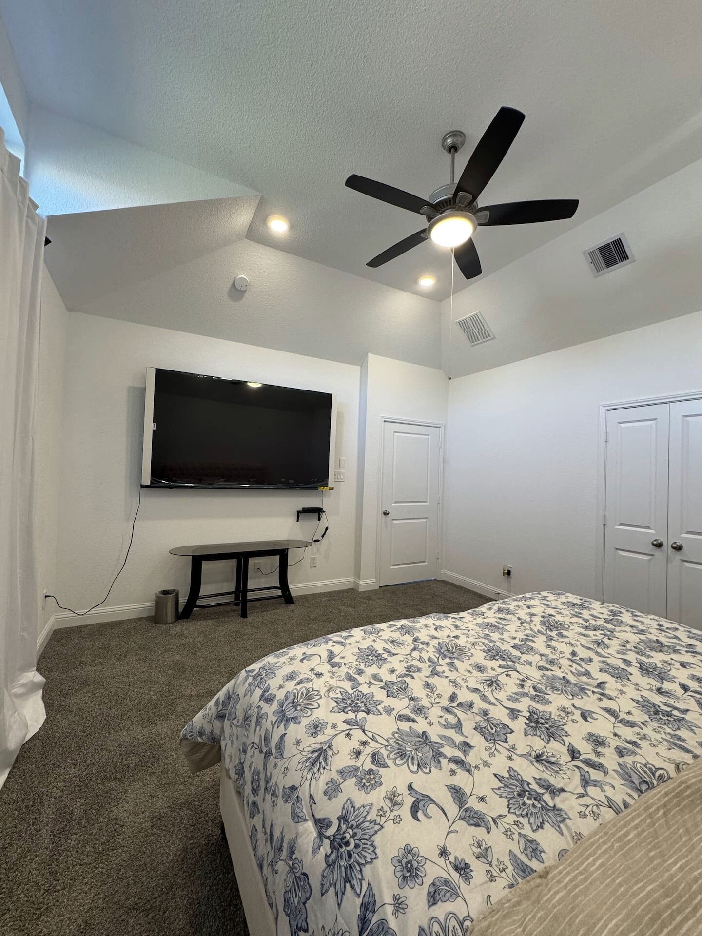 A bedroom with a bed, a ceiling fan, a TV mounted on the wall, and a door.