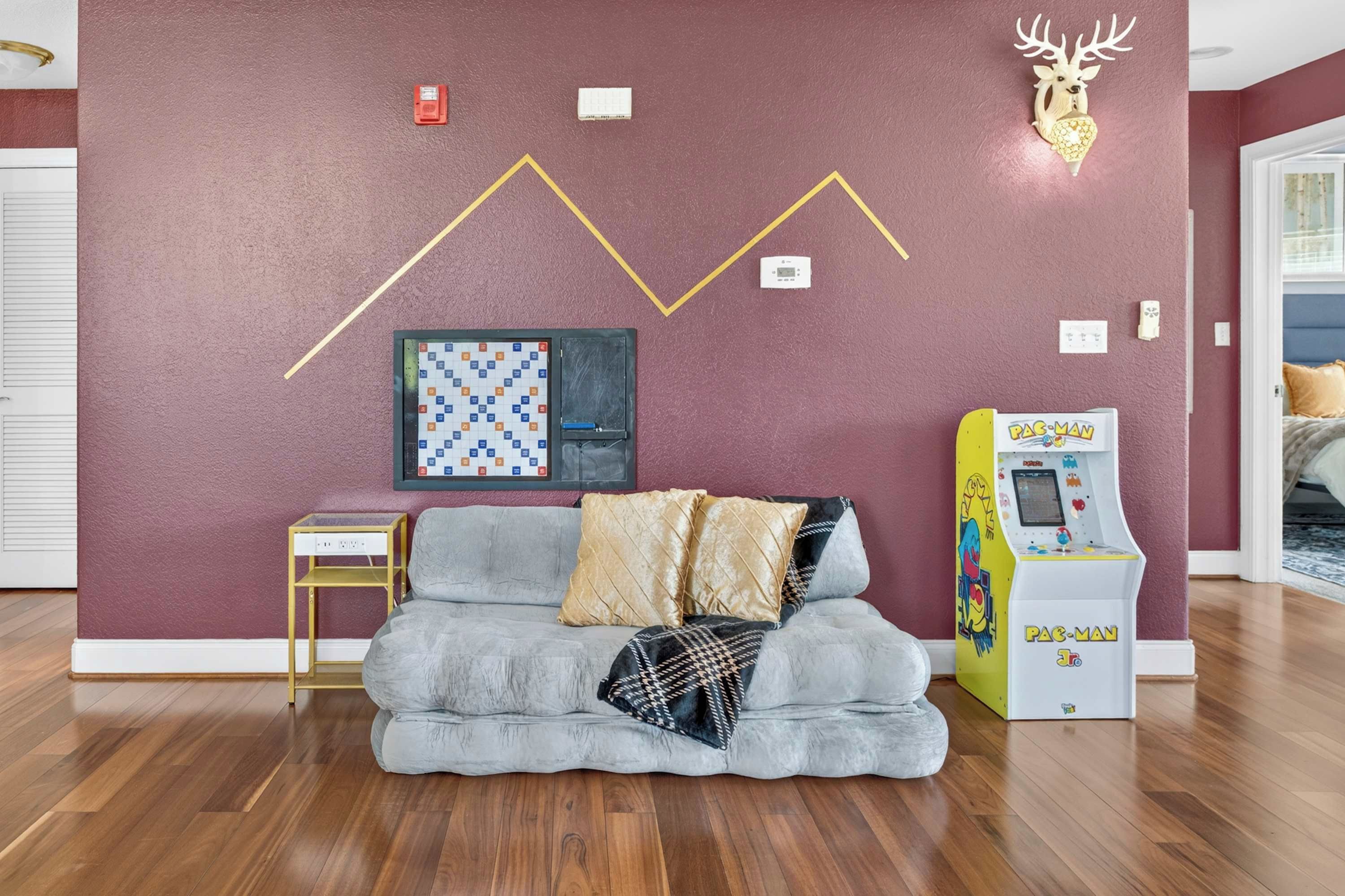 Una habitación con un sofá gris, una máquina arcade Pac-Man, un termostato montado en la pared y un juego de mesa enmarcado.