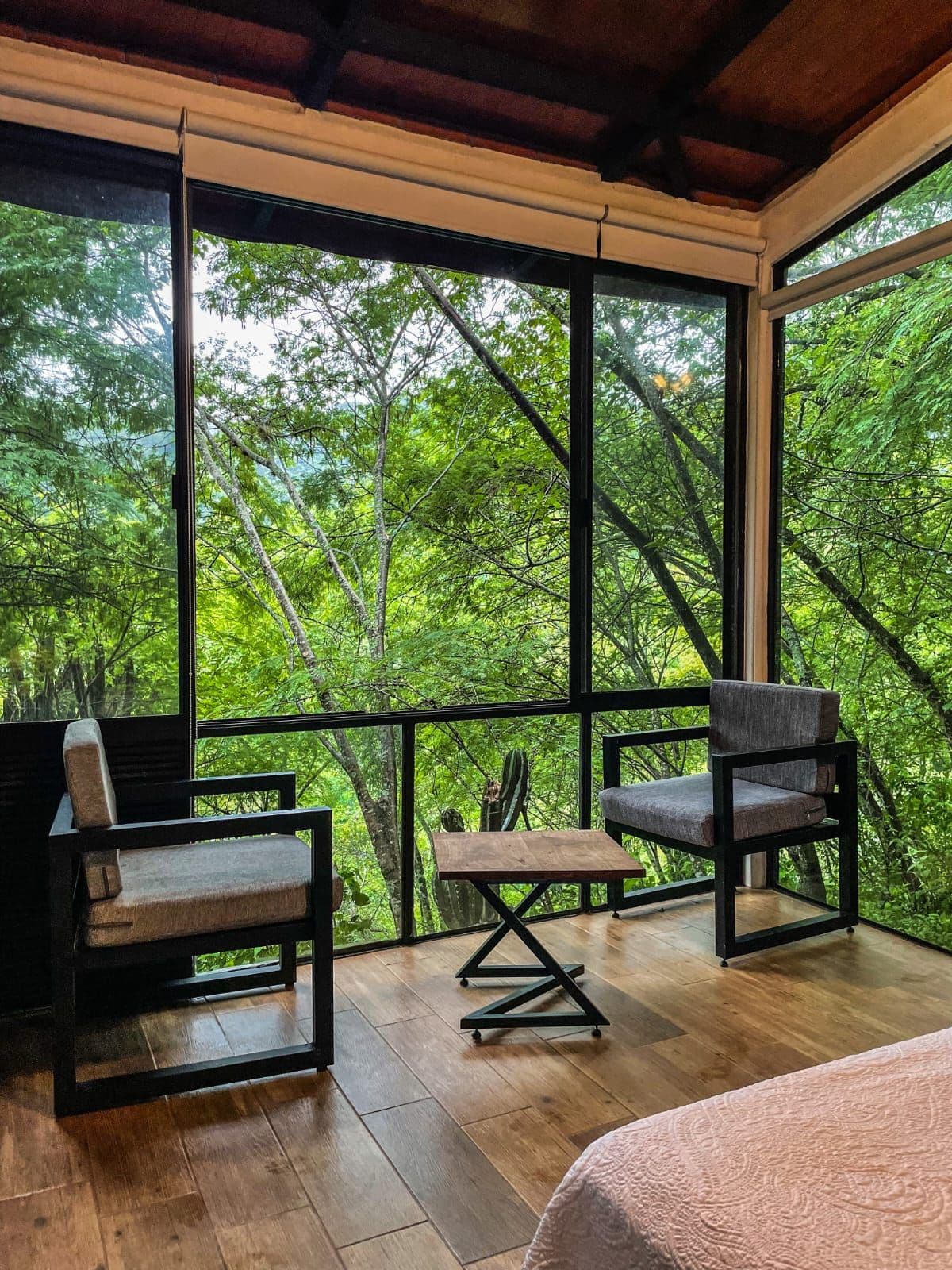 A room with a large window overlooking a forest, two chairs, a small table, and a bed with a textured blanket.