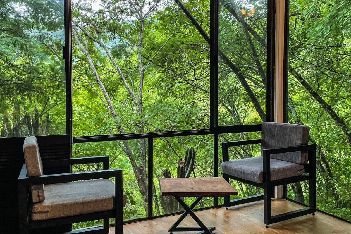 A room with a large window overlooking a forest, two chairs, a small table, and a bed with a textured blanket.