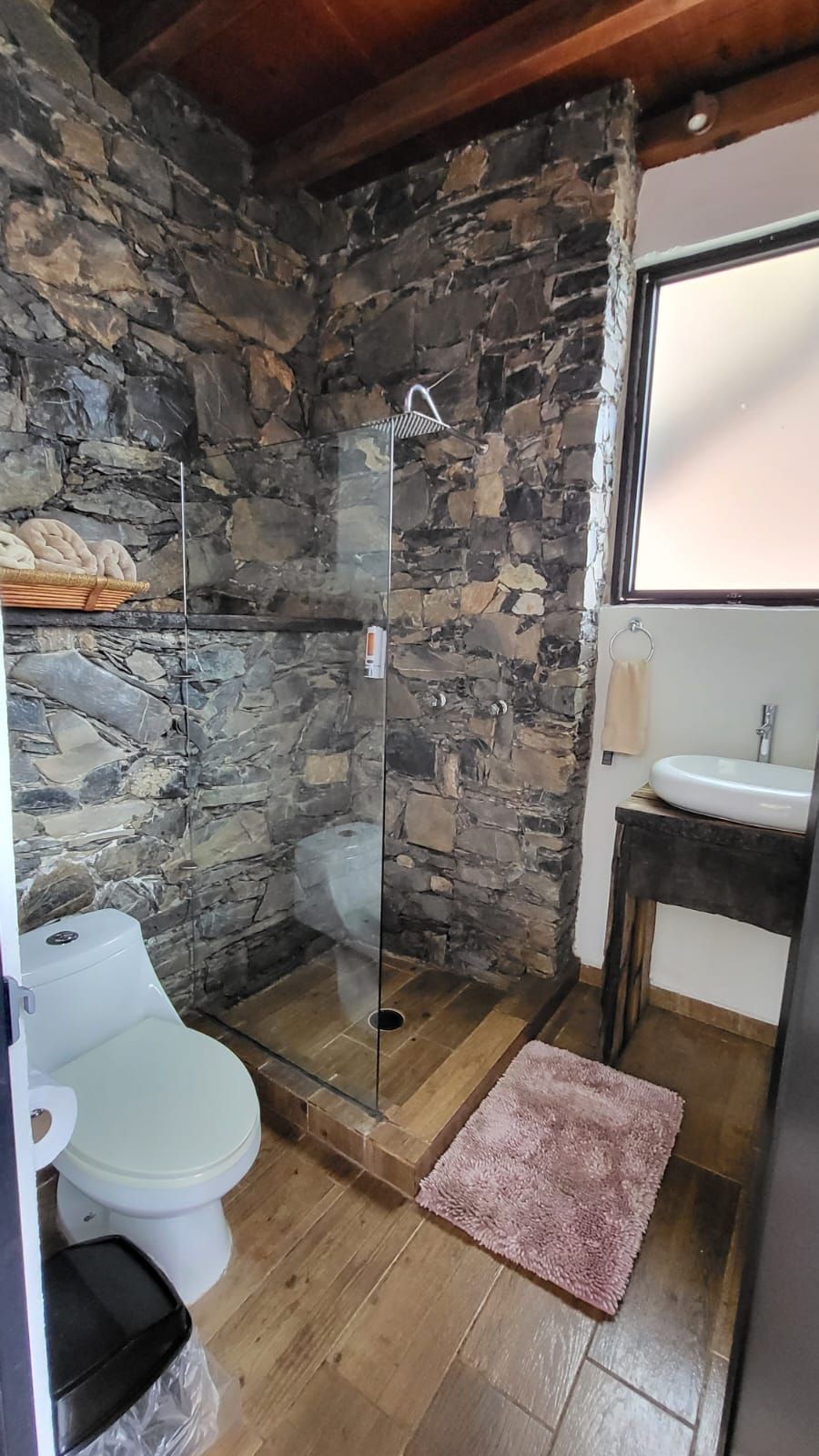 Un baño con pared de piedra, ducha, inodoro, lavabo y ventana.