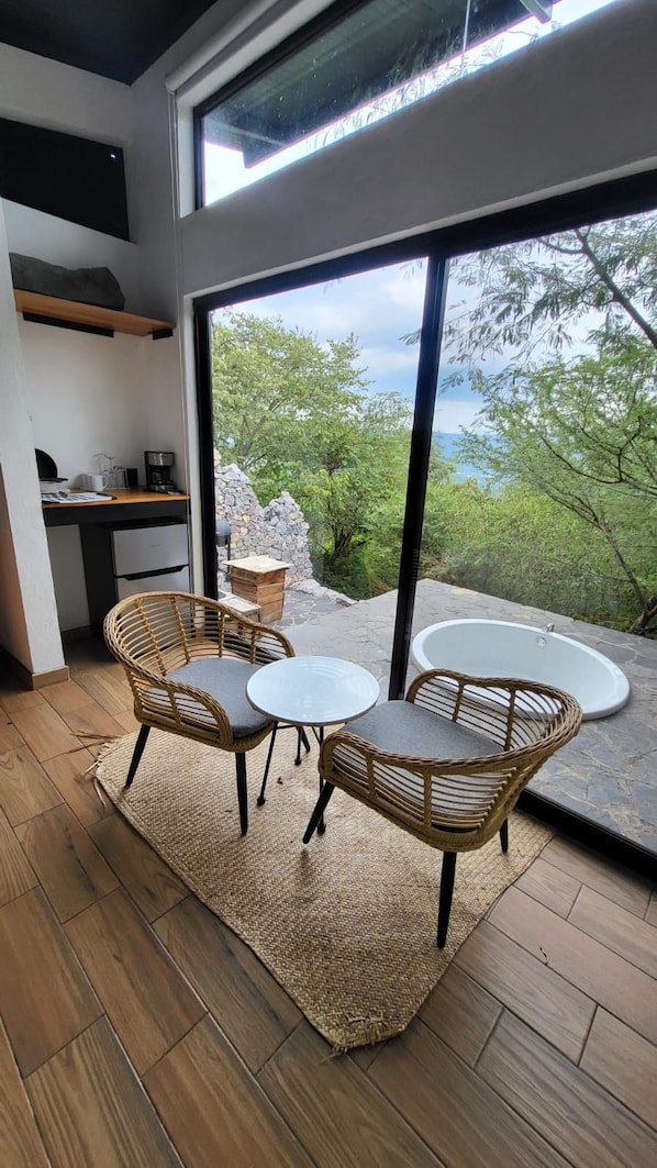 A modern room with a large window, a dining table, two wicker chairs, a small kitchen area, and a stone wall outside.