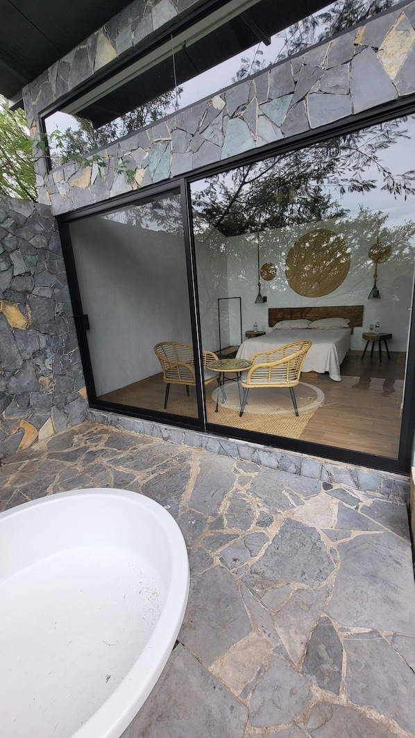 A modern bathroom with a stone wall, a large bathtub, and a glass-enclosed shower area with wicker chairs.