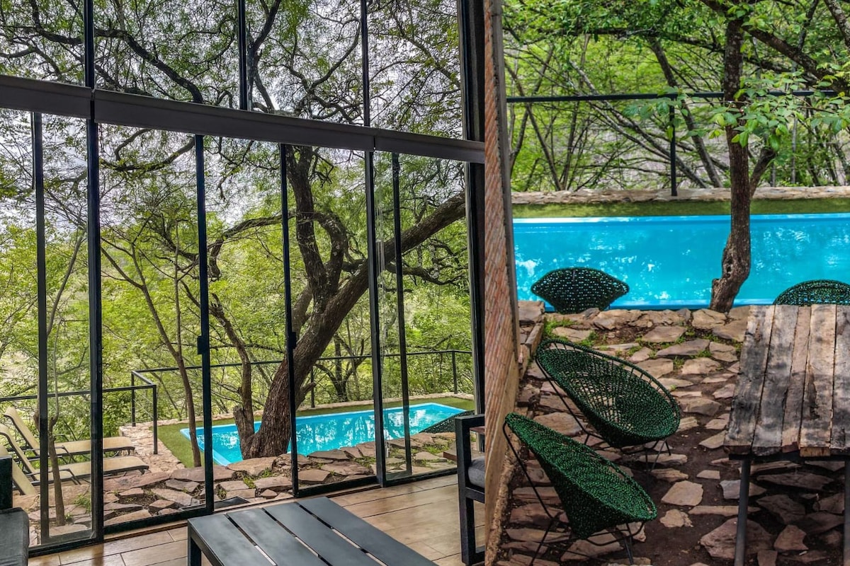 A poolside area with a view of a swimming pool, surrounded by trees and stone walls, and furnished with outdoor seating.