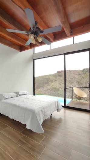 A bedroom with a bed, a ceiling fan, a large window, and a view of a landscape.