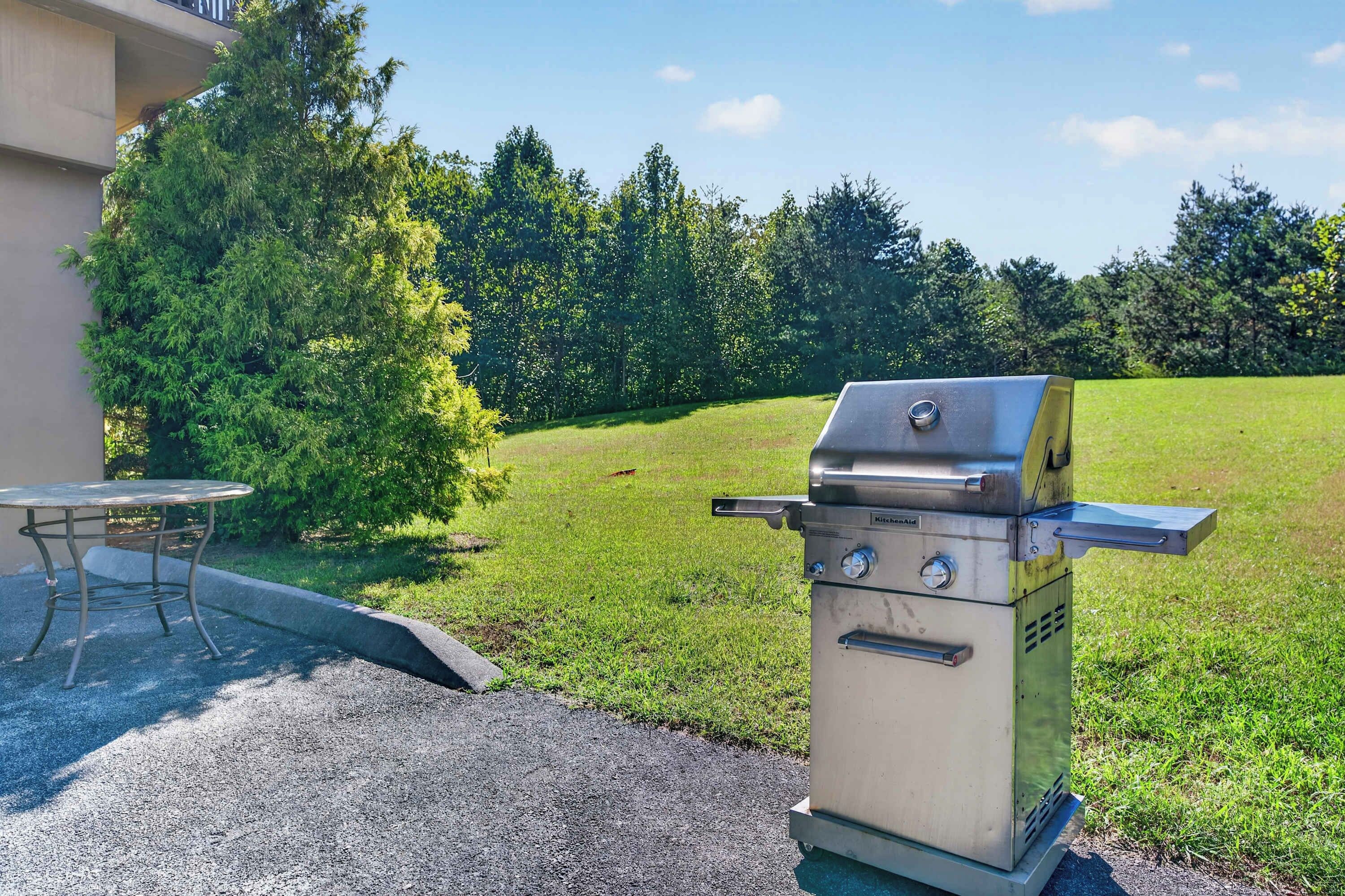 A portable gas grill with a control panel, a metal table, and a chair in a grassy outdoor area.