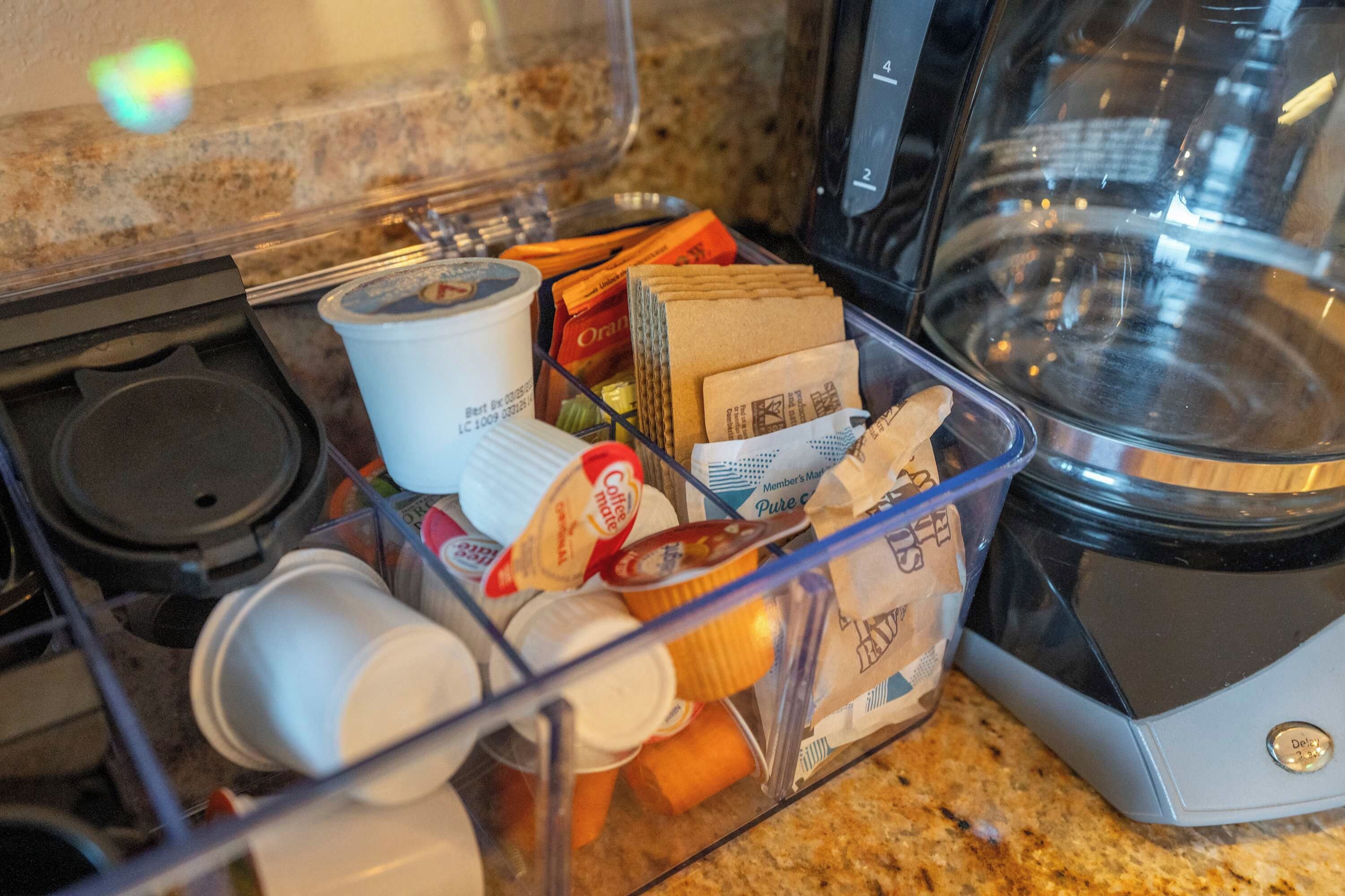 Un coin cuisine équipé d’une machine à café, d’un mixeur et d’un récipient en plastique contenant divers articles, notamment des capsules de café et un sachet de café moulu.
