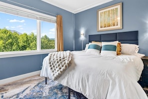 A bedroom with a large bed, a nightstand, a lamp, a window with a view of trees, and a painting on the wall. - Penthouse w/ Sauna, Arcade, Free Dollywood Tickets (Gatlinburg)