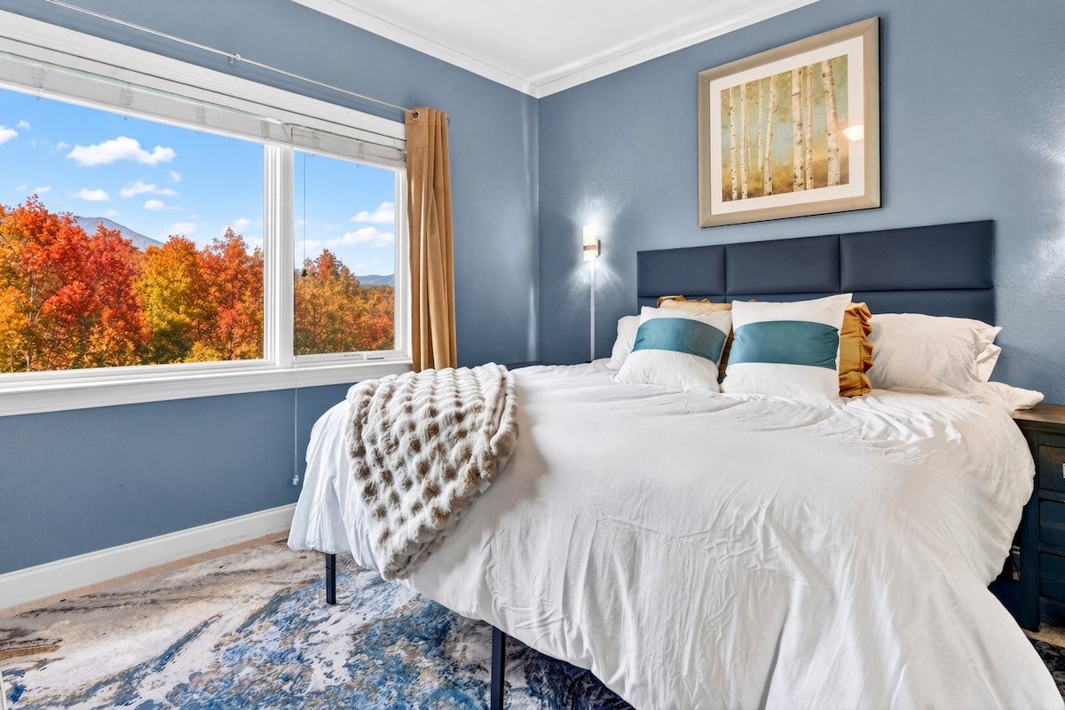 A bedroom with a large bed, a window with a view of autumn trees, a framed picture on the wall, and a nightstand.