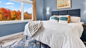 A bedroom with a large bed, a window with a view of autumn trees, a framed picture on the wall, and a nightstand. - Gatlinburg Condo: Sauna, Arcade, Spa, Incredible (Gatlinburg)
