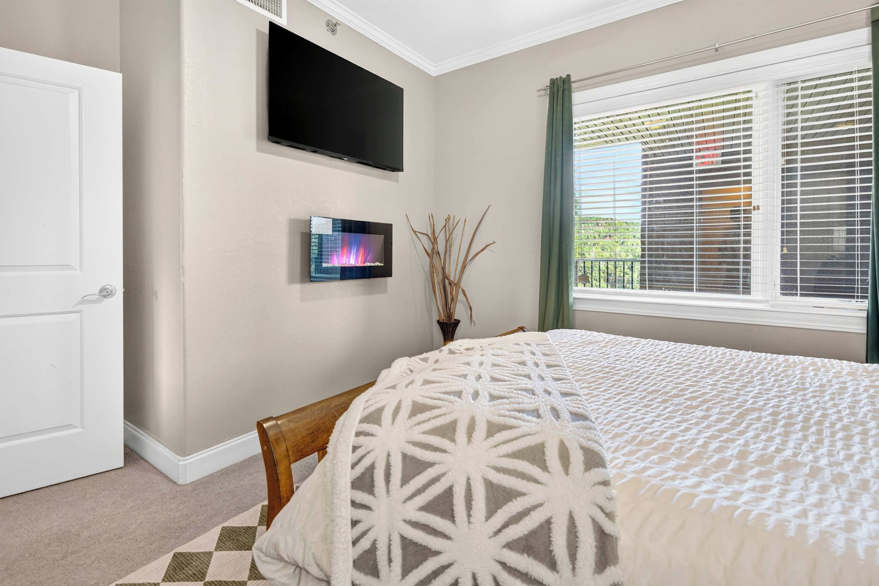A bedroom with a bed, a television mounted on the wall, a window with blinds, and a door.