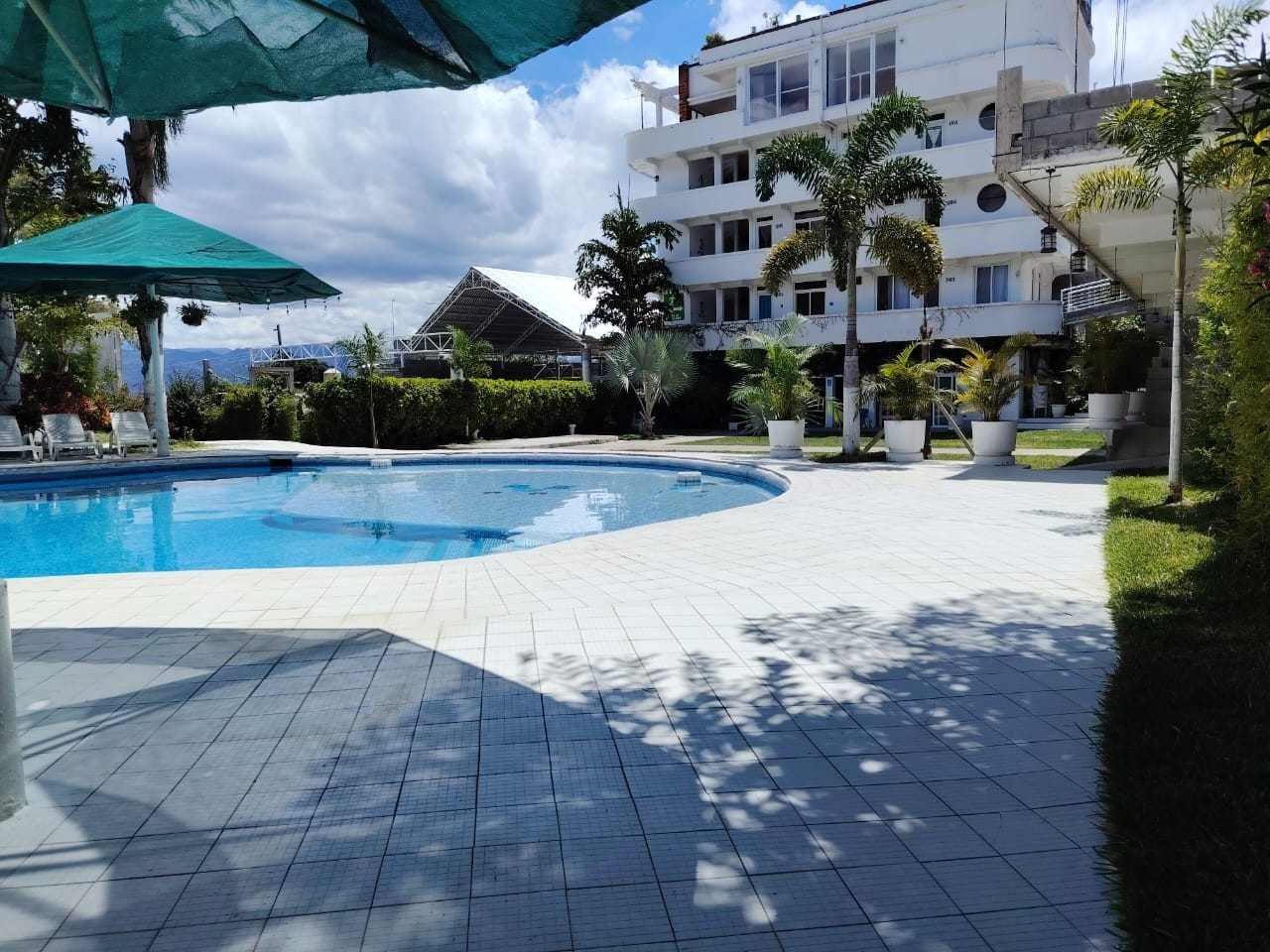 A swimming pool with a tiled deck, surrounded by greenery and a multi-story building.