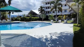 A swimming pool with a tiled deck, surrounded by greenery and a multi-story building.