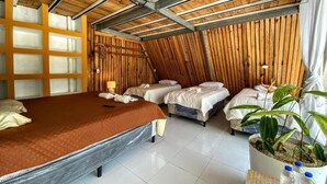 Ein Zimmer mit geneigter Decke, einem großen Bett, zwei kleineren Betten, einer Holzwandverkleidung, einer Topfpflanze und einem Deckenventilator.