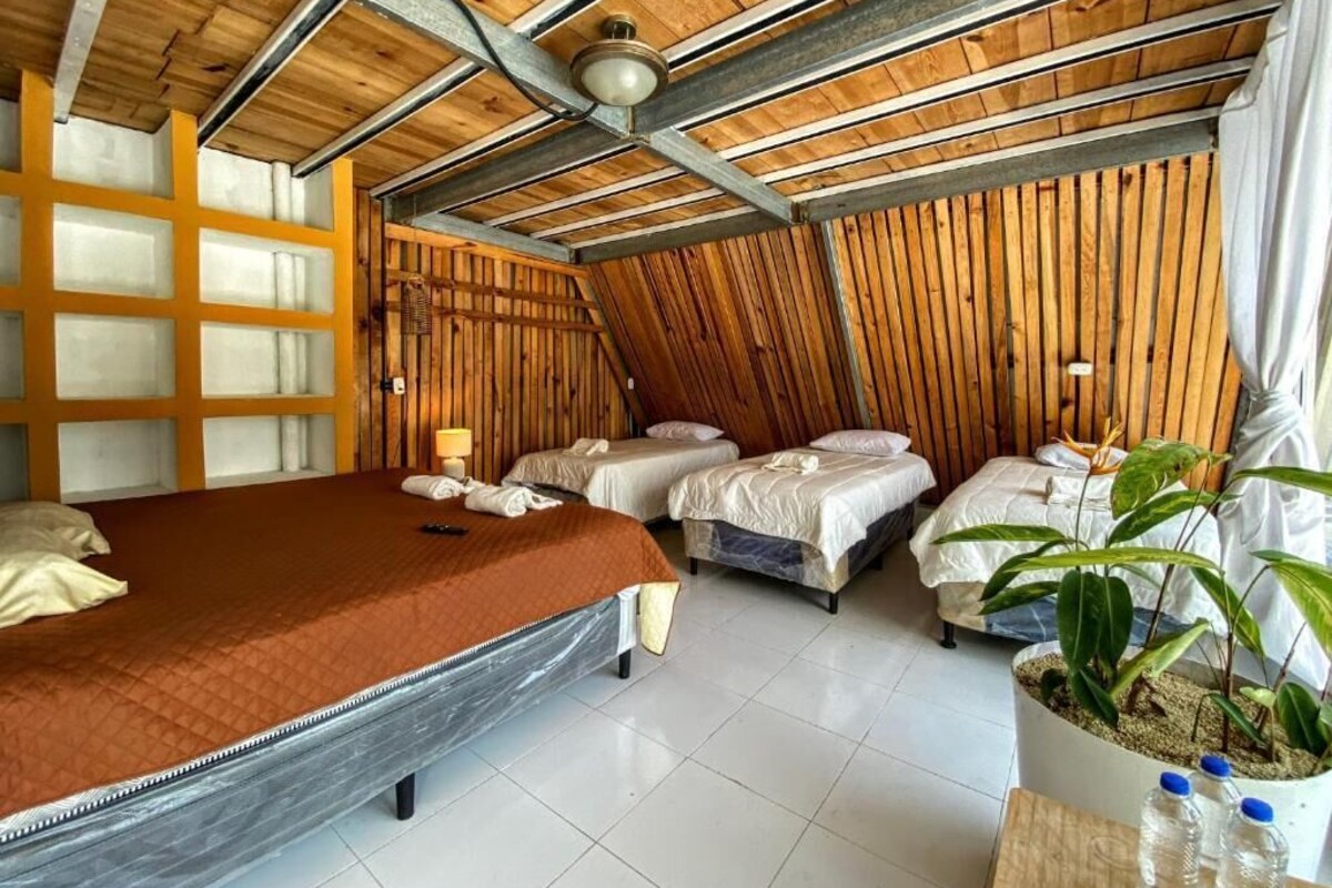 Una stanza con soffitto spiovente, un letto grande, due letti più piccoli, pannello murale in legno, una pianta in vaso e una ventola a soffitto.