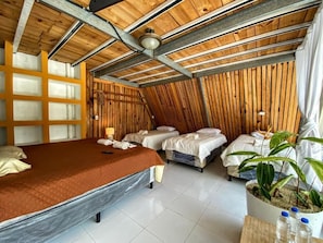 Una stanza con soffitto spiovente, un letto grande, due letti più piccoli, pannello murale in legno, una pianta in vaso e una ventola a soffitto.
