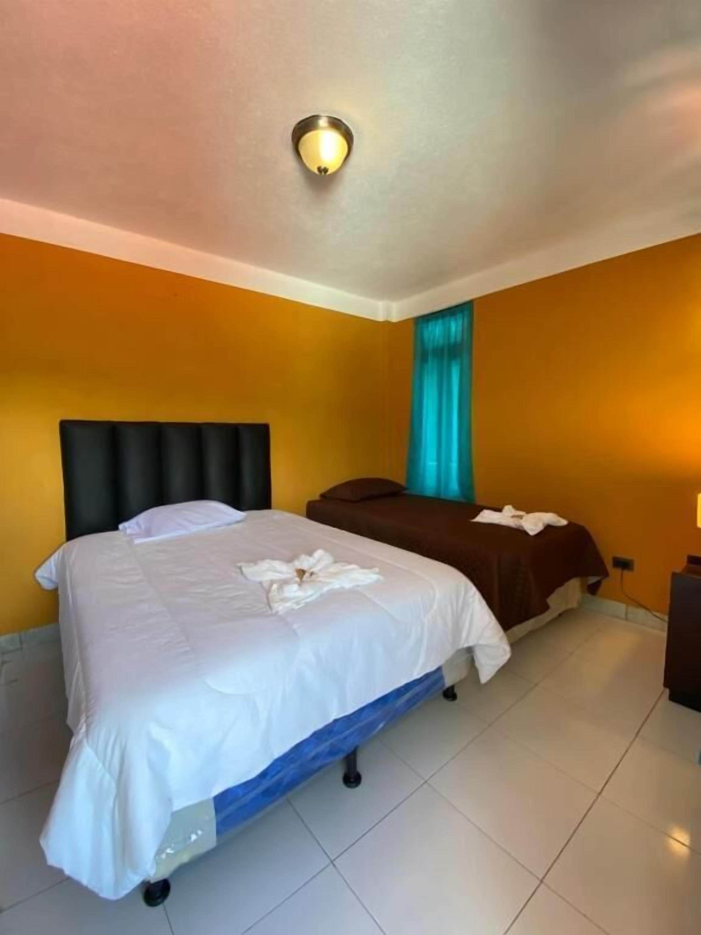 Habitación de hotel con cama, mesita de noche, ventana con cortinas y lámpara de pared.
