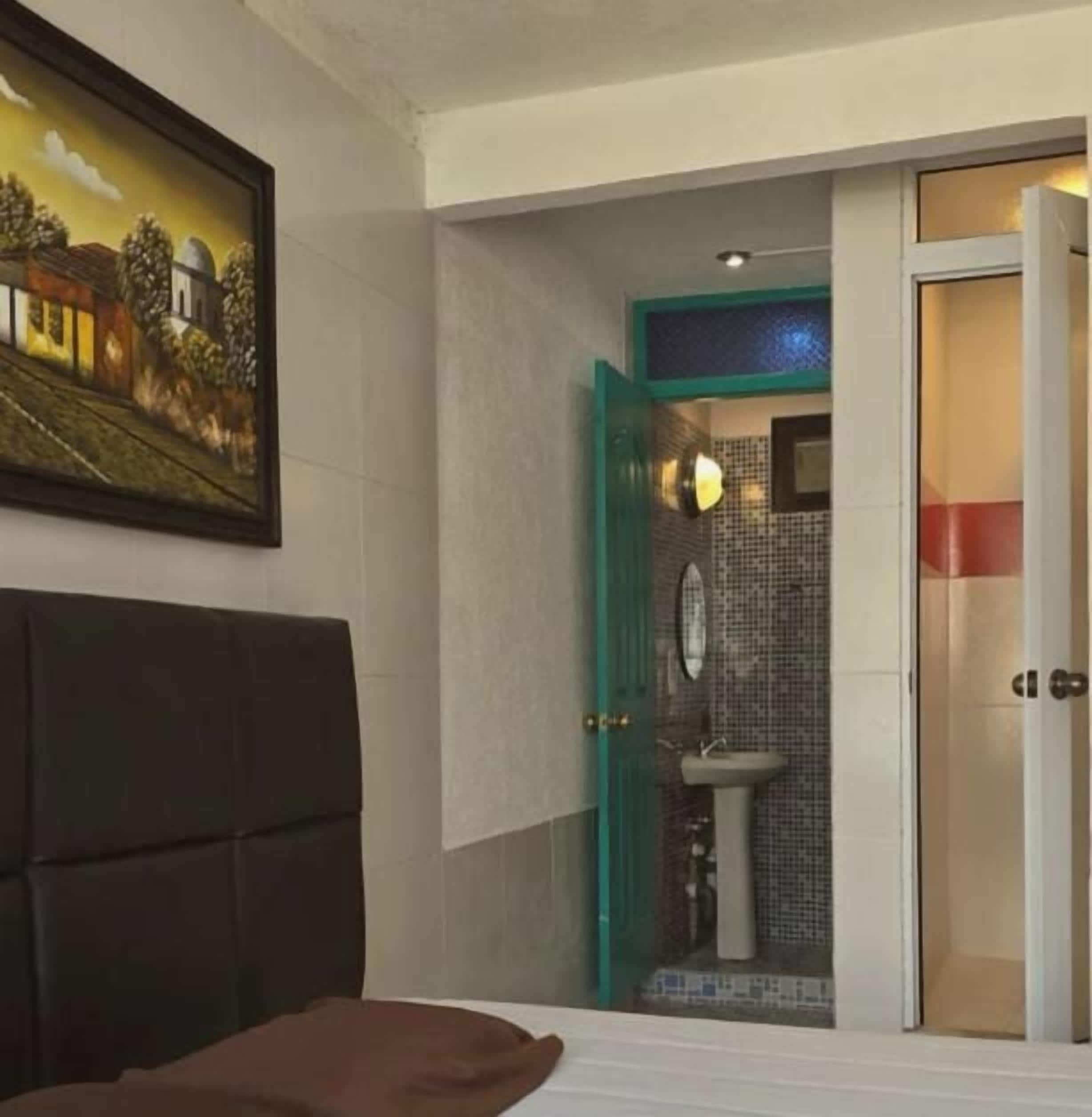 A hotel room with a bed, a painting on the wall, a bathroom with a sink and mirror, and a door leading to another room.